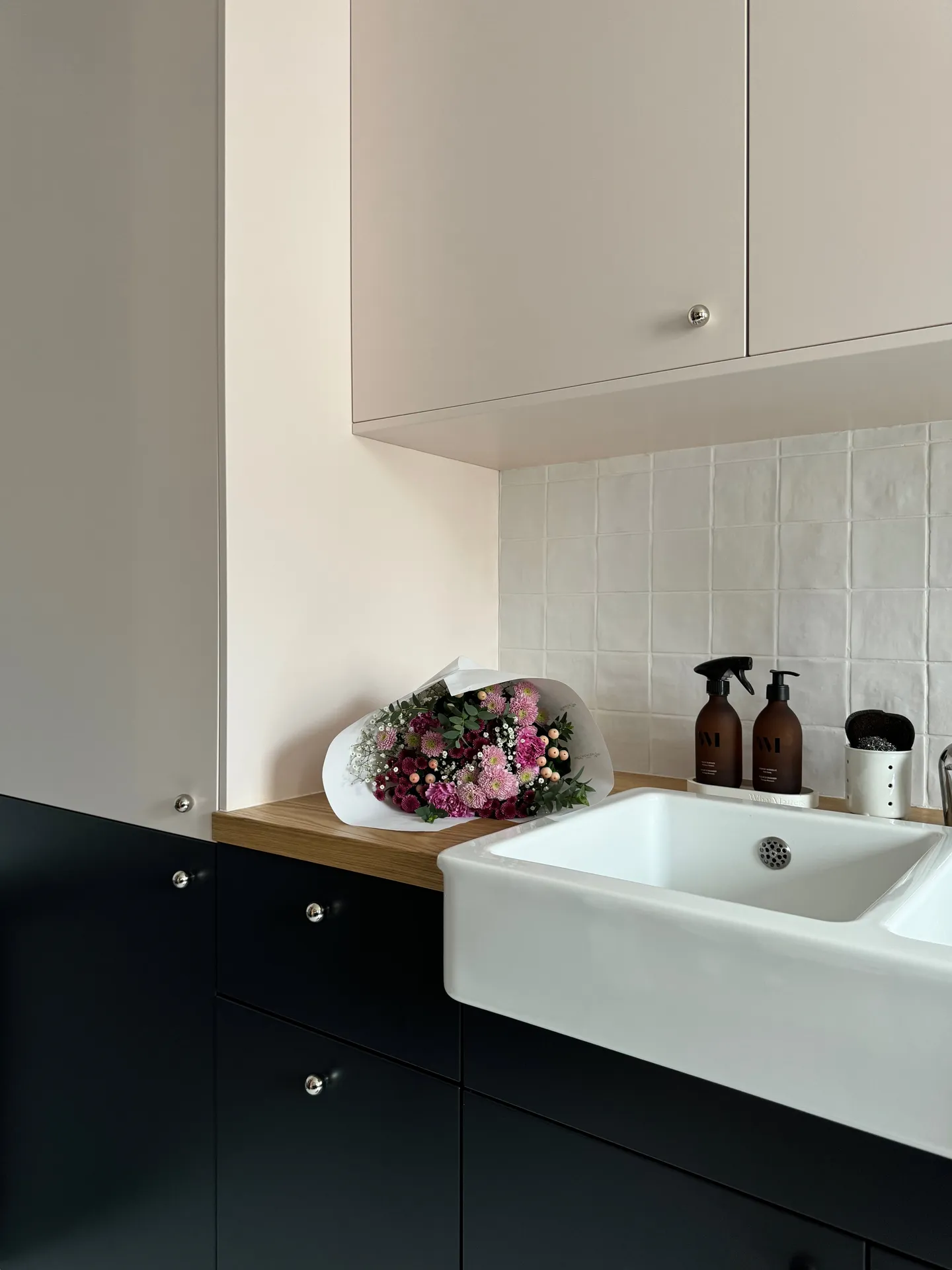 Studio Madame : architecte d’intérieur, Paris et Montmorency
Projet réalisé à Enghien-les-Bains.
Photographie d’intérieur : cuisine habillée de façades rose pâle et bleu nuit et composée d’un plan de travail en chêne, d’un évier en céramique avec mitigeur chromé, d’une faïence blanche artisanale pour la crédence et de poignées chromées en forme de bulle. Des bouteilles de produit ménager, un pot en porcelaine ainsi qu’un bouquet de fleurs dans des tons de rose sont posés sur le plan de travail.