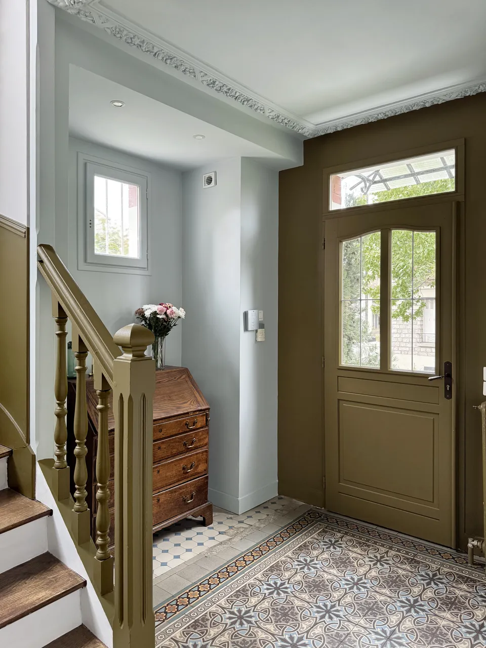 Studio Madame : architecte d’intérieur, Paris et Enghien-les-Bains.
Projet réalisé à Deuil-la-Barre.
Photographie d’intérieur : entrée d'une ancienne meulière rénovée à Deuil-la-Barre avec escalier ancien, moulures, plafond sculpté et niche bleu pâle, porte d’entrée vert bronze et carreaux de ciment d’origine au sol.