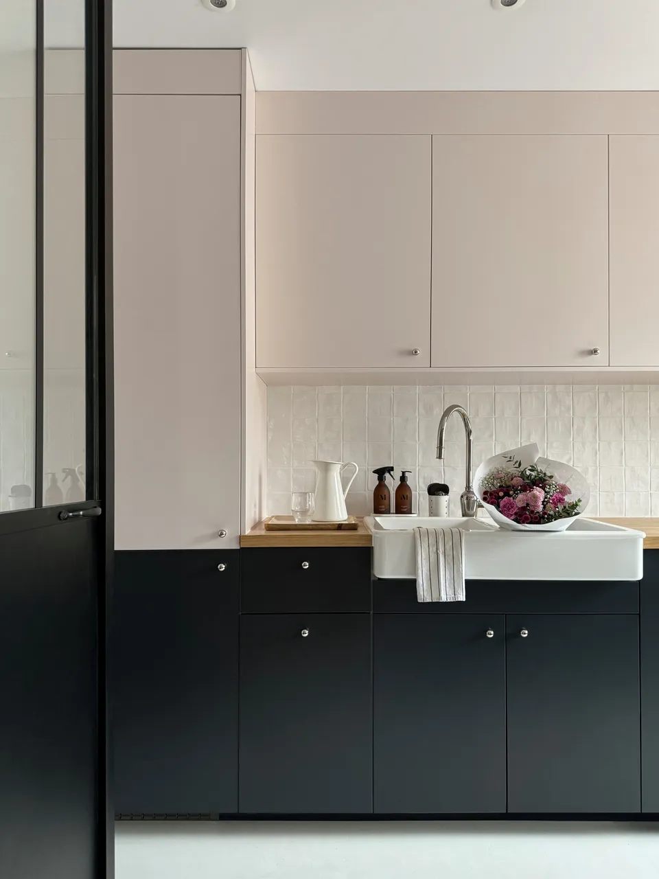 Studio Madame : architecte d’intérieur, Paris et Montmorency
Projet réalisé à Enghien-les-Bains.
Photographie d’intérieur : cuisine habillée de façades rose pâle et bleu nuit et composée d’un plan de travail en chêne, d’un évier en céramique avec mitigeur chromé, d’une faïence blanche artisanale pour la crédence, de poignées chromées en forme de bulle et d’un sol en béton ciré gris clair. Des bouteilles de produit ménager, un pot en porcelaine, un plateau avec une carafe ancienne blanche et un verre, ainsi qu’un bouquet de fleurs dans des tons de rose sont posés sur le plan de travail et dans l’évier. Un torchon sèche sur le bord de l’évier. La porte permettant d’entrer dans la cuisine est une verrière industrielle noire.